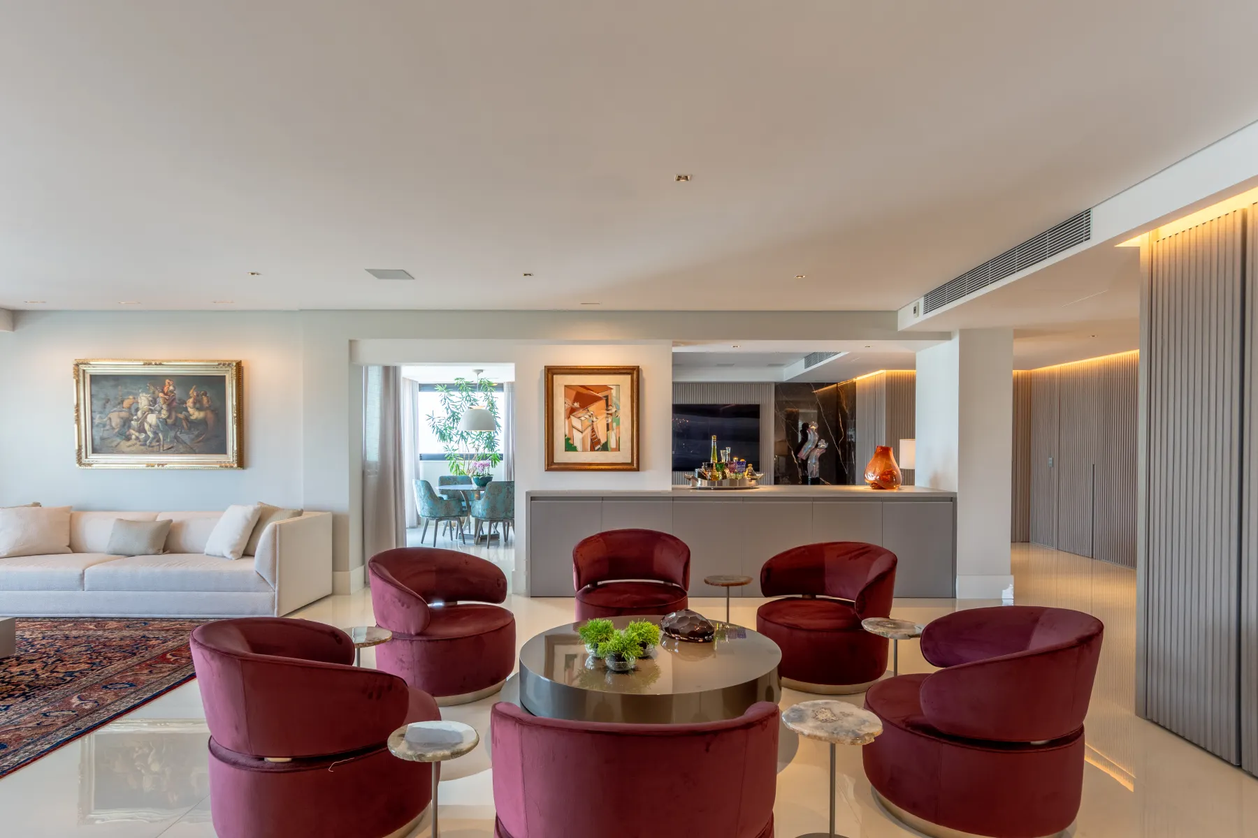 Sala de jantar com poltronas de veludo borgonha ao redor de mesa redonda, tapete persa e quadros na parede